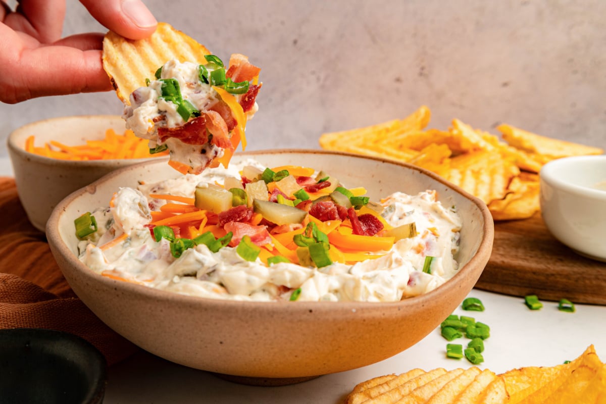 A hand dips a ridged potato chip into a bowl of loaded dip topped with shredded cheese, bacon, and green onions. More chips and a small white bowl are in the background.