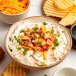 A bowl of creamy dip topped with shredded cheddar, chopped bacon, and green onions, surrounded by ridged potato chips and a bowl of shredded cheese.