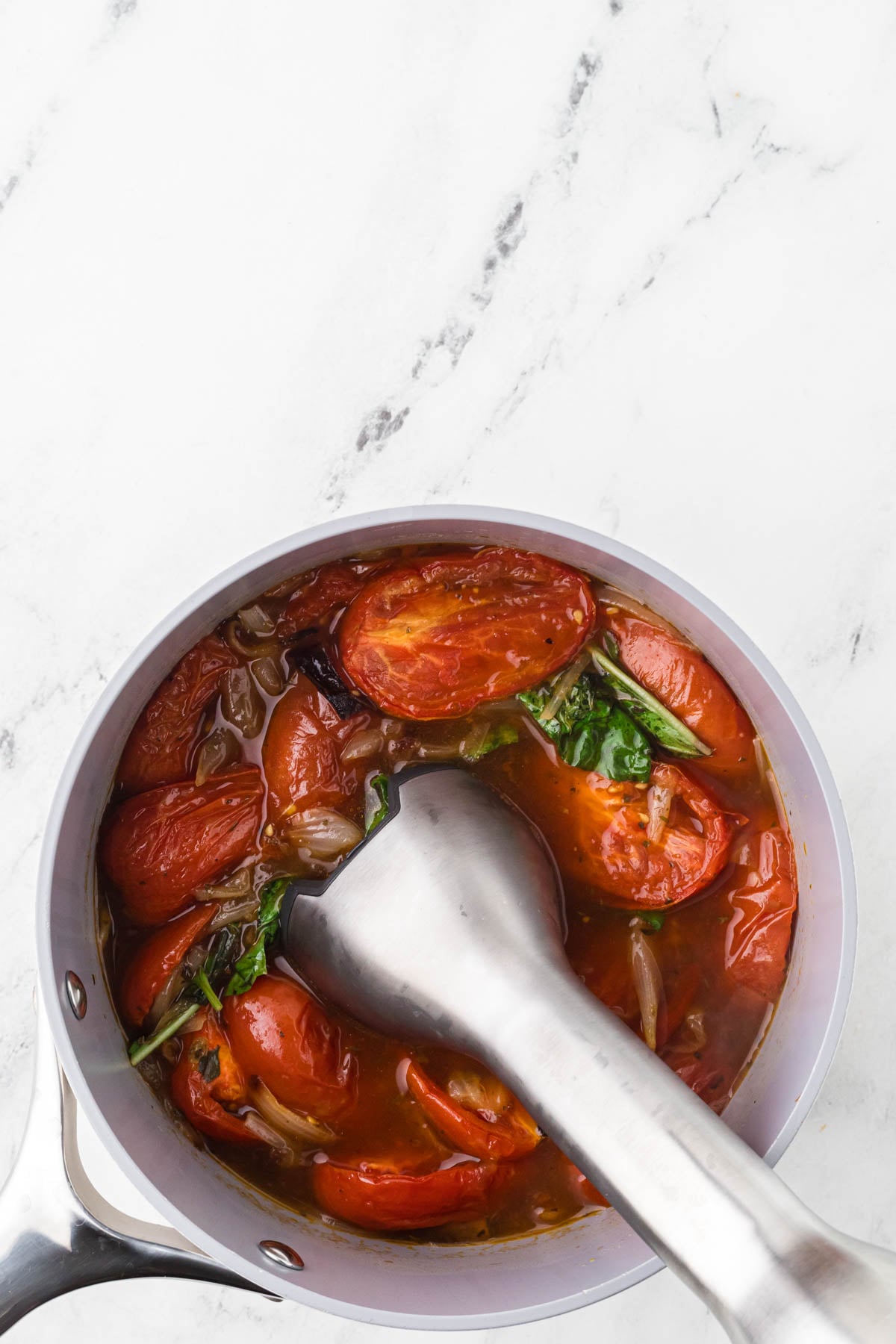 A saucepan with cooked tomatoes, onions, and herbs being blended with an immersion blender on a white marble countertop.