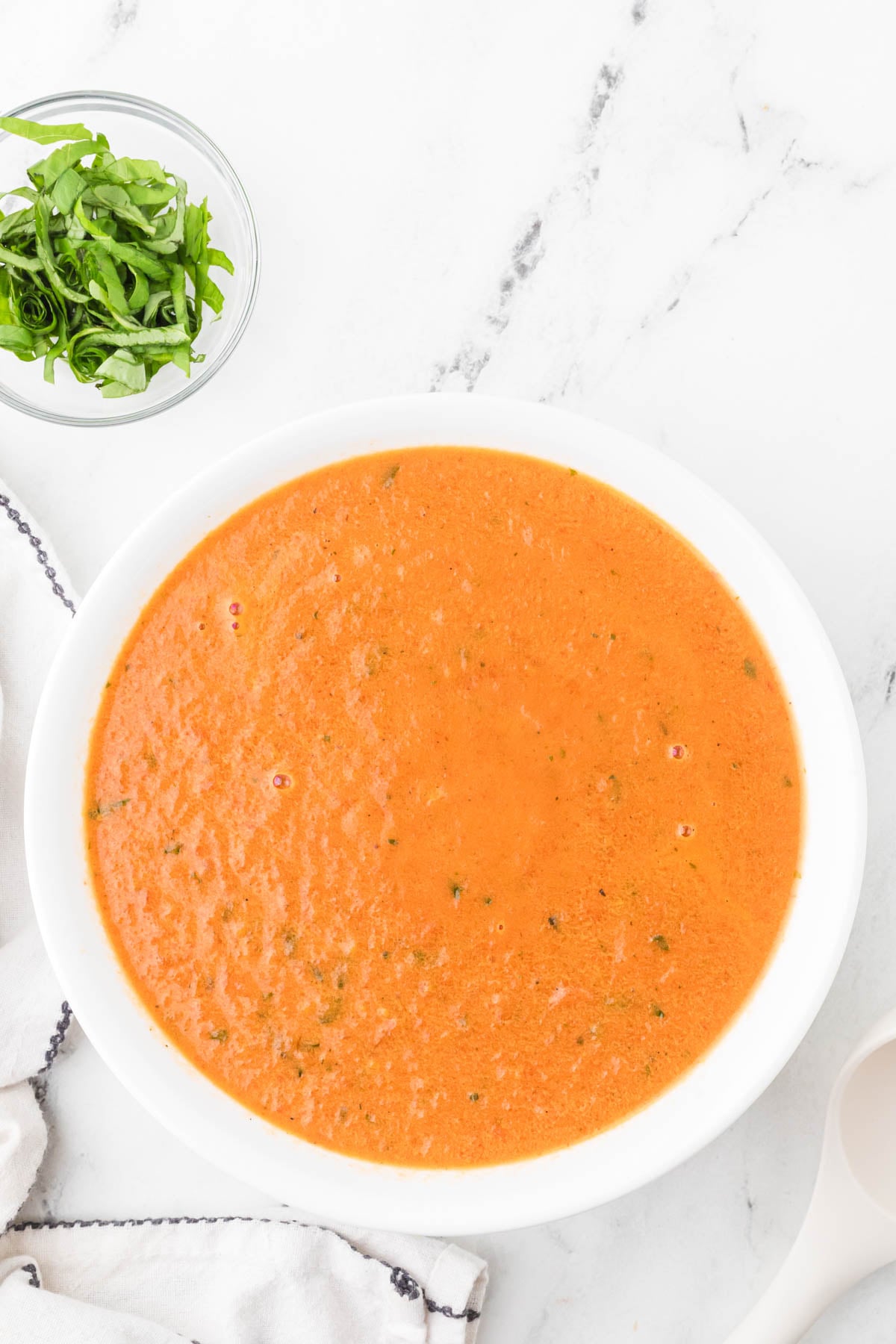A bowl of creamy tomato soup sits on a white marble surface, with a small dish of chopped green herbs nearby.