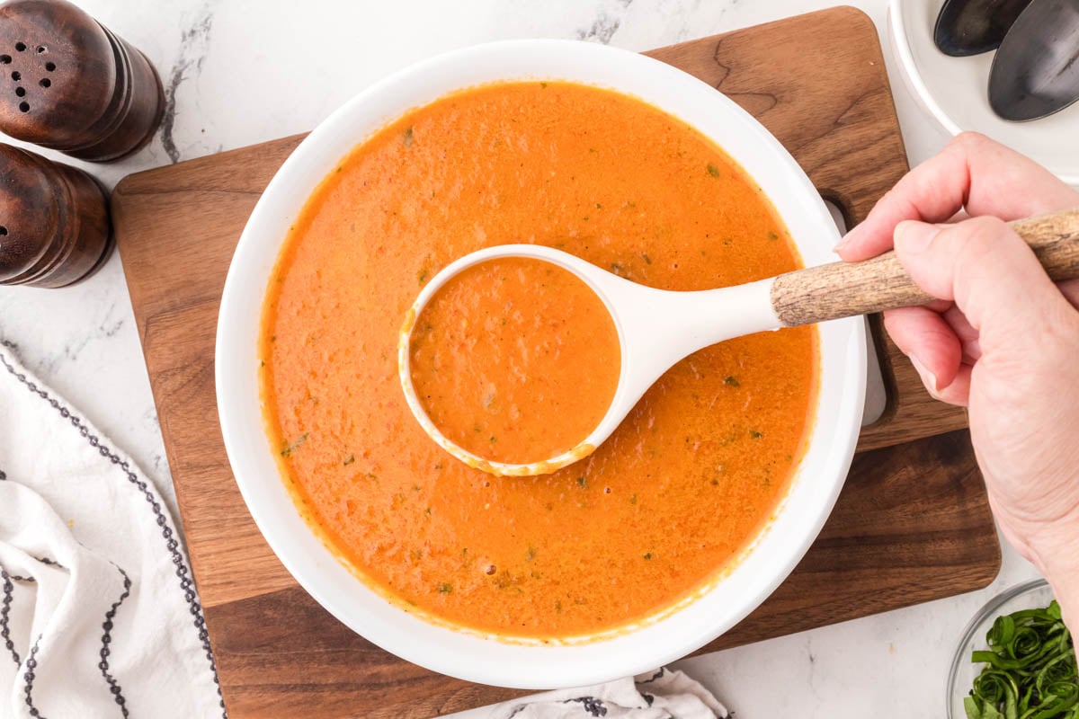 A hand holds a ladle above a bowl of tomato soup on a wooden board, with salt and pepper shakers and a cloth napkin nearby.