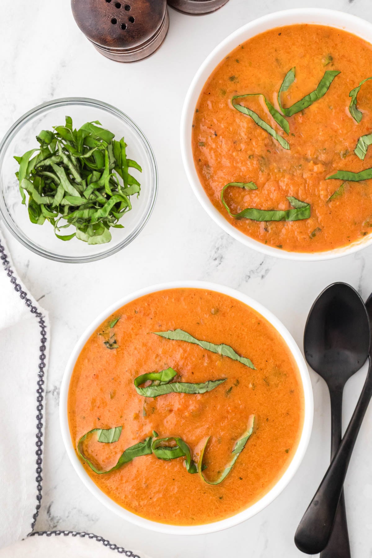 Two bowls of tomato soup topped with sliced basil, next to a small bowl of fresh basil, black spoons, a napkin, and a pair of salt and pepper shakers on a marble surface.