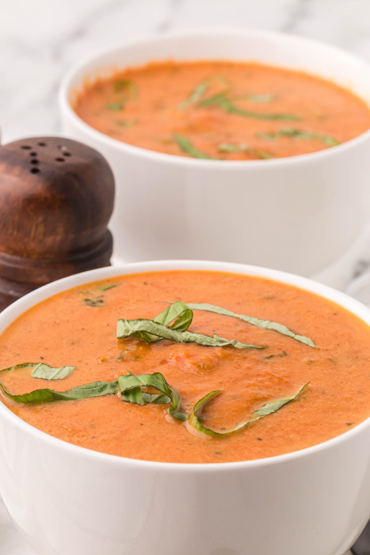 Two white bowls filled with creamy tomato soup, garnished with fresh basil strips, sit on a white surface beside a wooden pepper grinder.