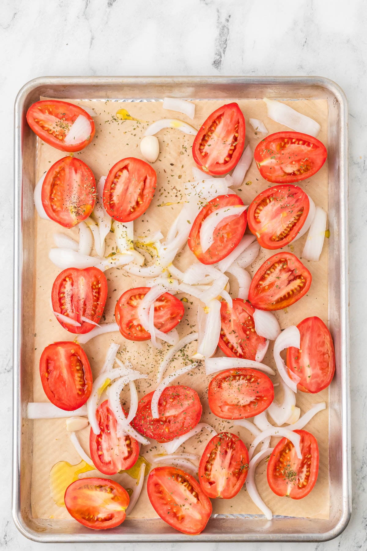 Halved tomatoes, sliced onions, whole garlic cloves, and herbs arranged on a parchment-lined baking sheet, drizzled with oil.