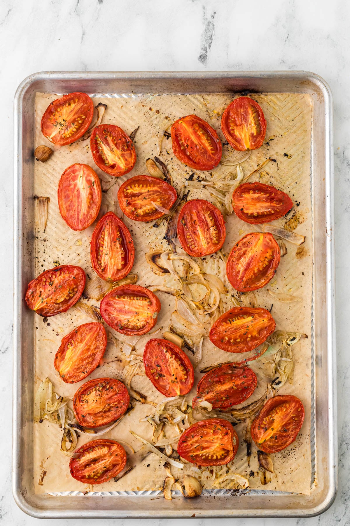 Halved roasted tomatoes and sliced onions on a parchment-lined baking sheet, lightly charred and seasoned, on a marble countertop.
