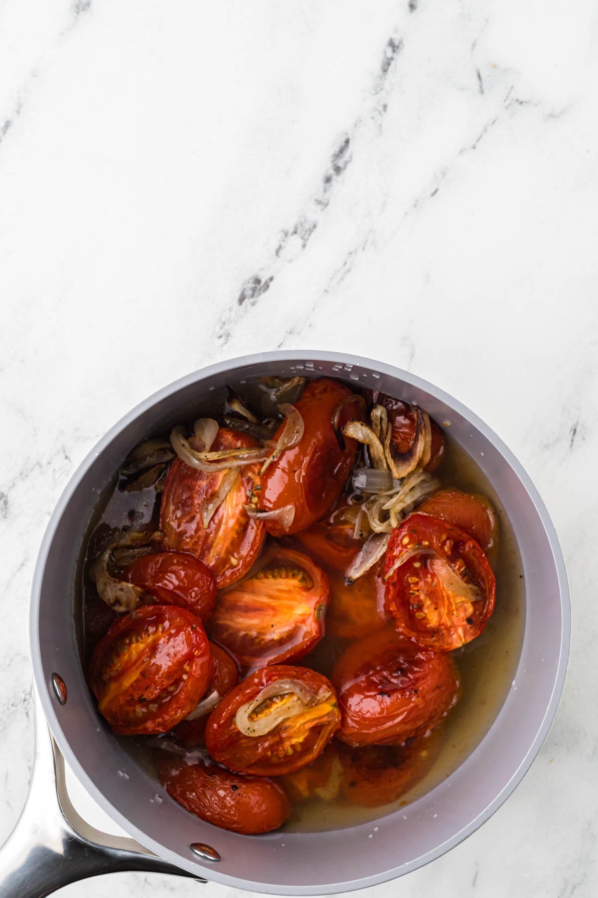 A saucepan on a marble surface contains halved tomatoes, sliced onions, and liquid, appearing to simmer.
