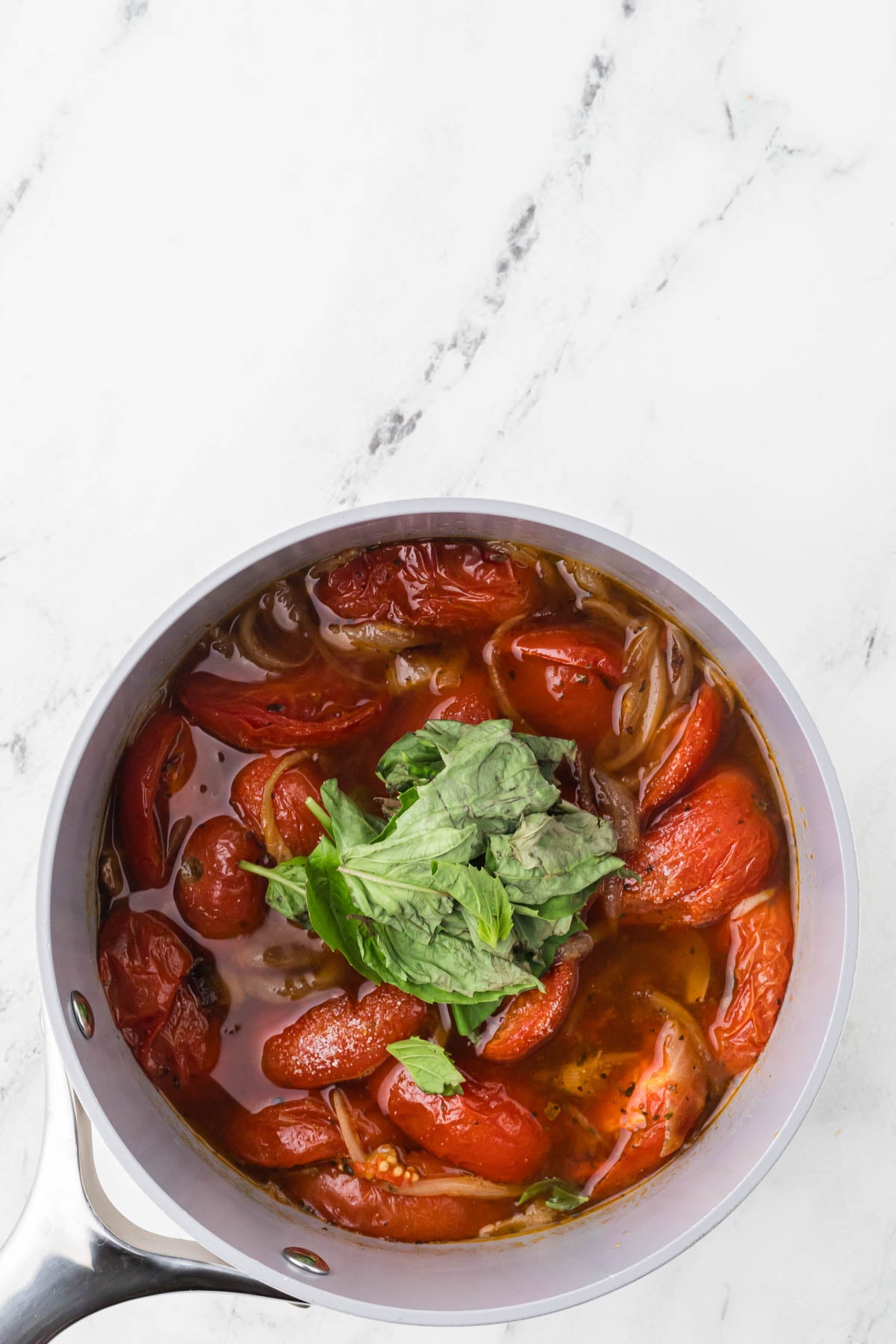 Top-down view of a light gray saucepan filled with blistered cherry tomatoes, sautéed onions, and fresh green basil, resting on a white marble surface.