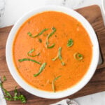 A white bowl filled with creamy orange tomato soup, garnished with thin strips of fresh green basil, sits on a wooden board.
