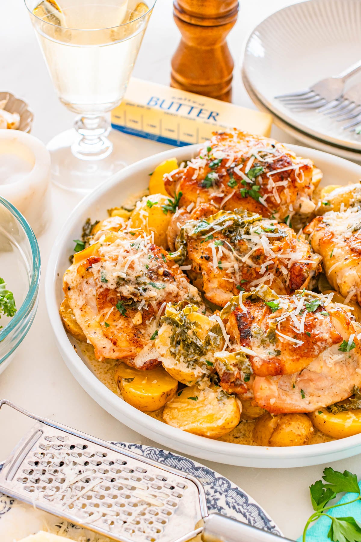 Roasted chicken thighs on a plate with yellow potatoes, topped with herbs and shredded cheese, next to a glass of white wine and a stick of butter.