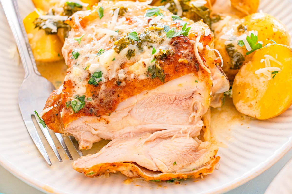 A plate with sliced roasted chicken topped with herbs and cheese, served with halved yellow potatoes. A fork is placed on the plate.