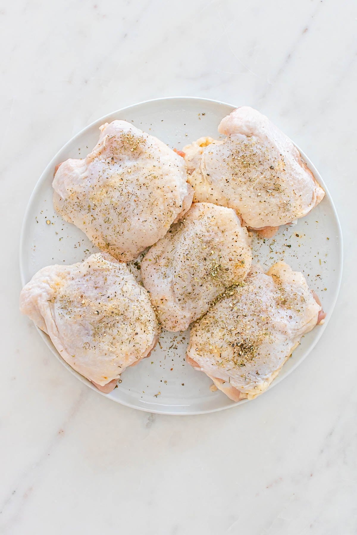 Five raw, skin-on chicken thighs arranged on a white plate, sprinkled with dried herbs and spices, set on a light-colored marble surface.