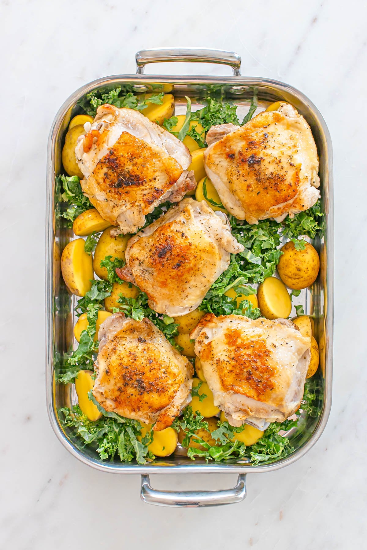 Five baked chicken thighs on a bed of sliced yellow potatoes and kale in a rectangular metal roasting pan on a marble surface.