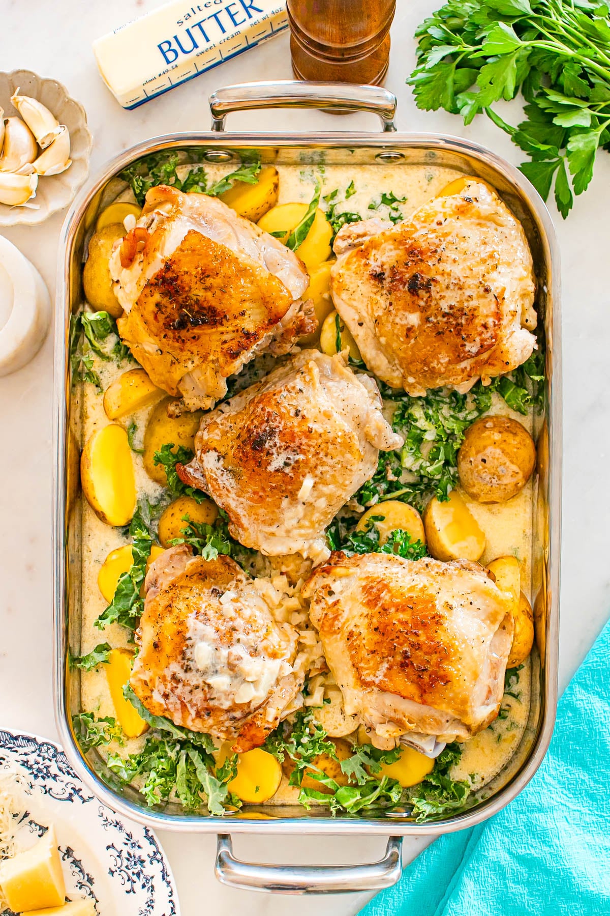 Baked chicken thighs on a bed of sliced potatoes and greens in a rectangular pan, surrounded by butter, garlic, parsley, and a blue napkin.
