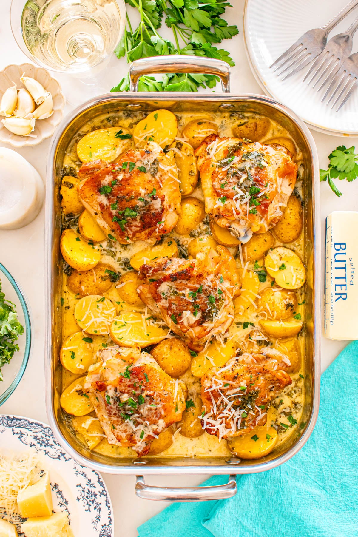 Roasted chicken thighs with golden potatoes in a creamy sauce, garnished with herbs and cheese, served in a baking dish on a table set with utensils, parsley, butter, and a glass of wine.