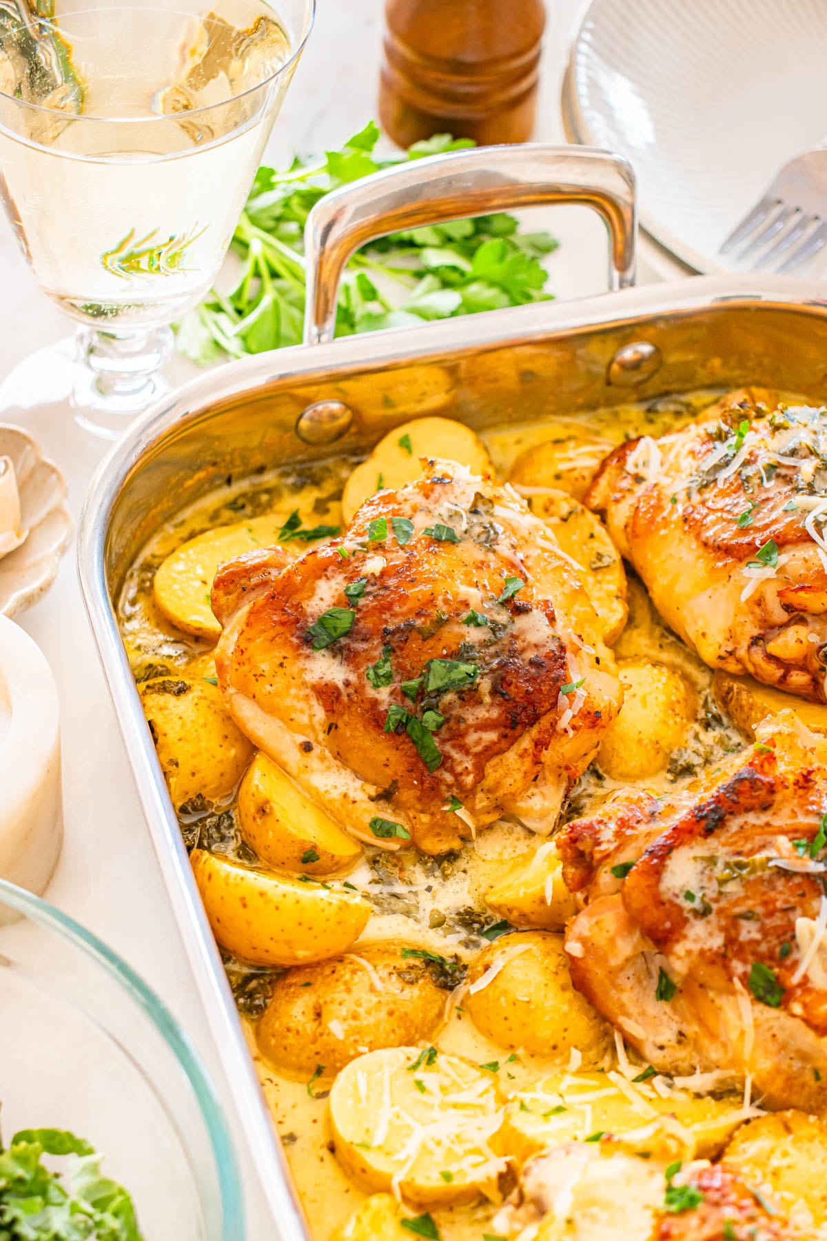 Roasted chicken thighs with potatoes, garnished with fresh herbs and grated cheese, served in a metal baking dish on a table with a glass of white wine and parsley in the background.
