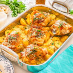 Baked chicken thighs and halved yellow potatoes topped with herbs and grated cheese in a metal roasting pan on a table.