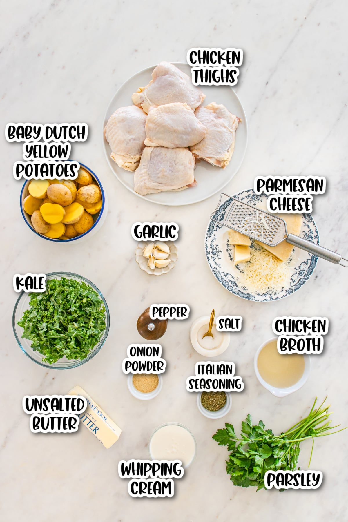 Top-down view of ingredients for a chicken dish, including chicken thighs, potatoes, kale, garlic, cheese, spices, butter, cream, broth, and parsley, arranged on a marble surface.