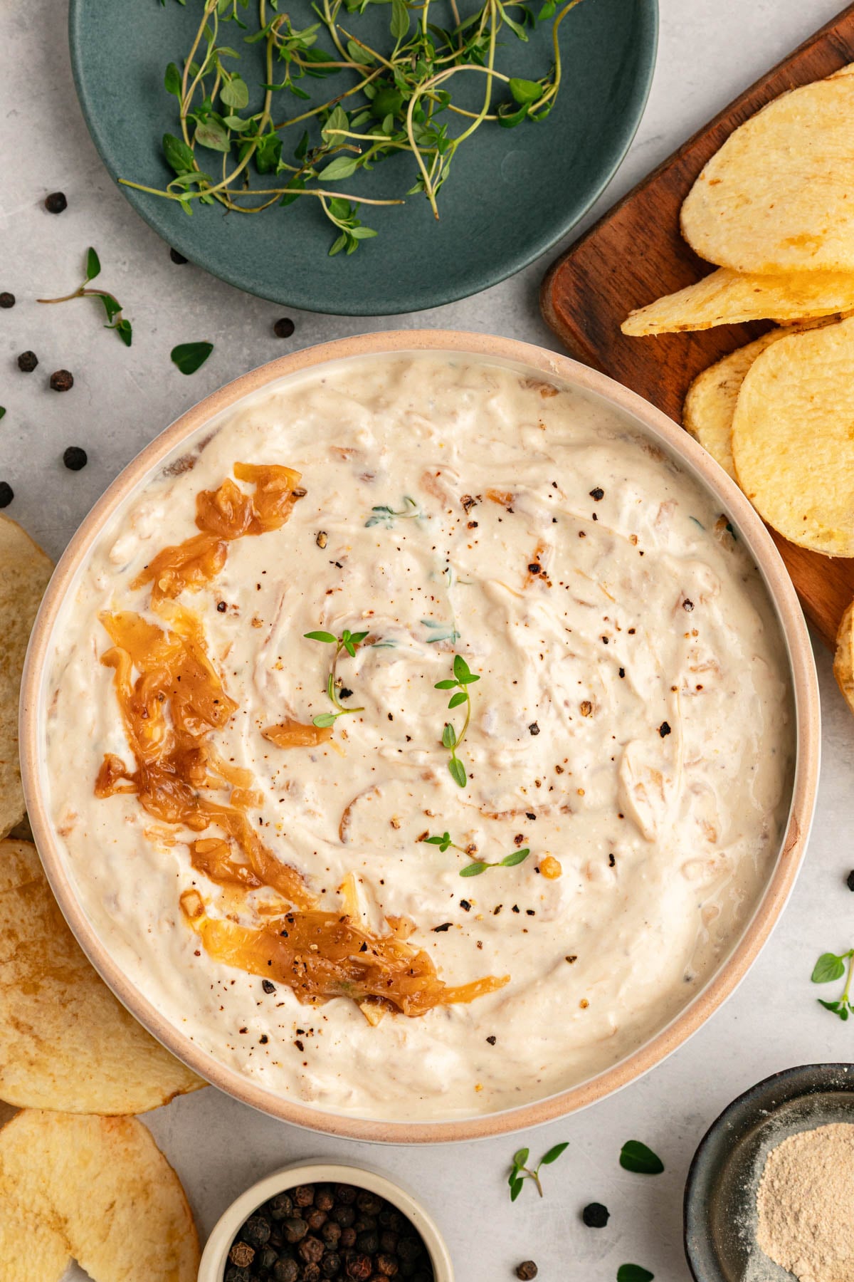 A bowl of creamy dip garnished with caramelized onions and herbs, surrounded by potato chips, black peppercorns, and fresh thyme on a table.