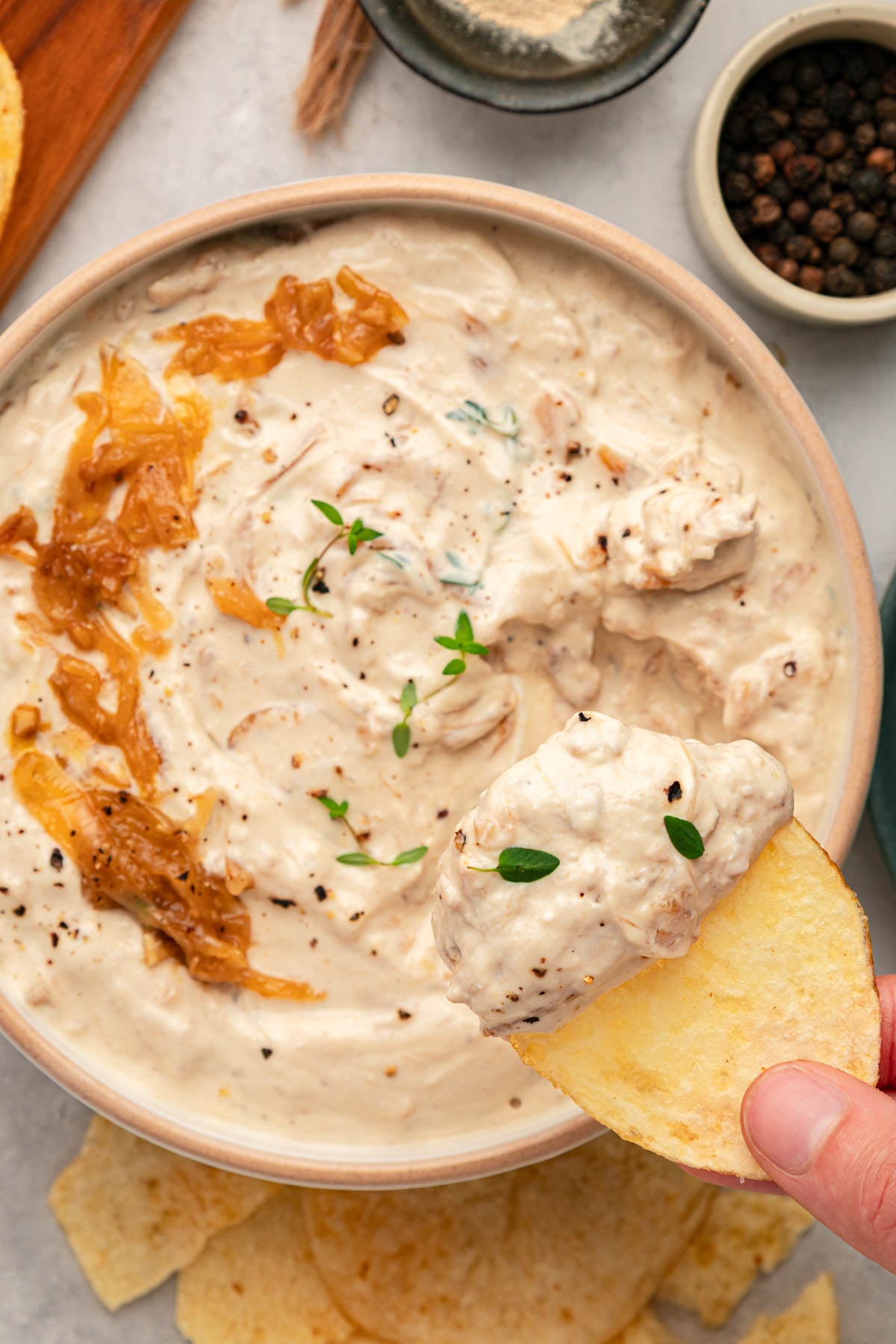 A hand holds a potato chip dipped in creamy onion dip, garnished with herbs and black pepper; caramelized onions are visible in the dip.