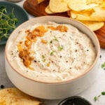 A bowl of creamy dip topped with caramelized onions and herbs, with potato chips and greens nearby.