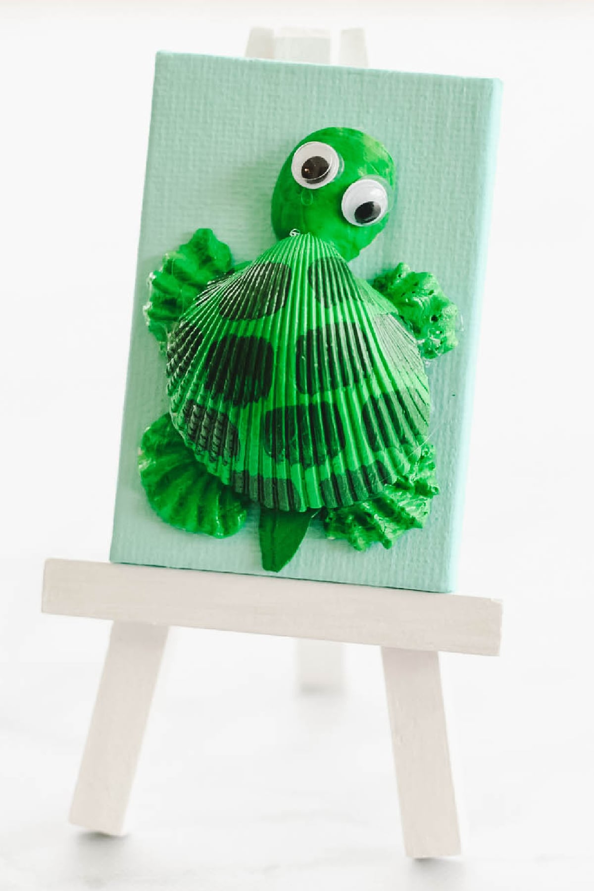 A small canvas on an easel displays a turtle crafted from a green-painted seashell and other materials, with googly eyes and green paint details.