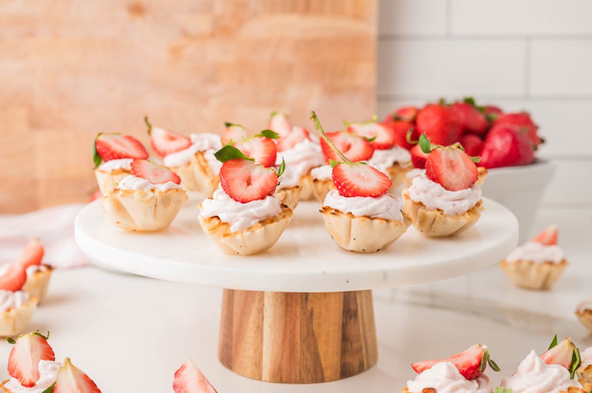 Strawberry cheesecake bites topped with strawberry halves displayed on a white cake stand, with fresh strawberries in a bowl in the background.