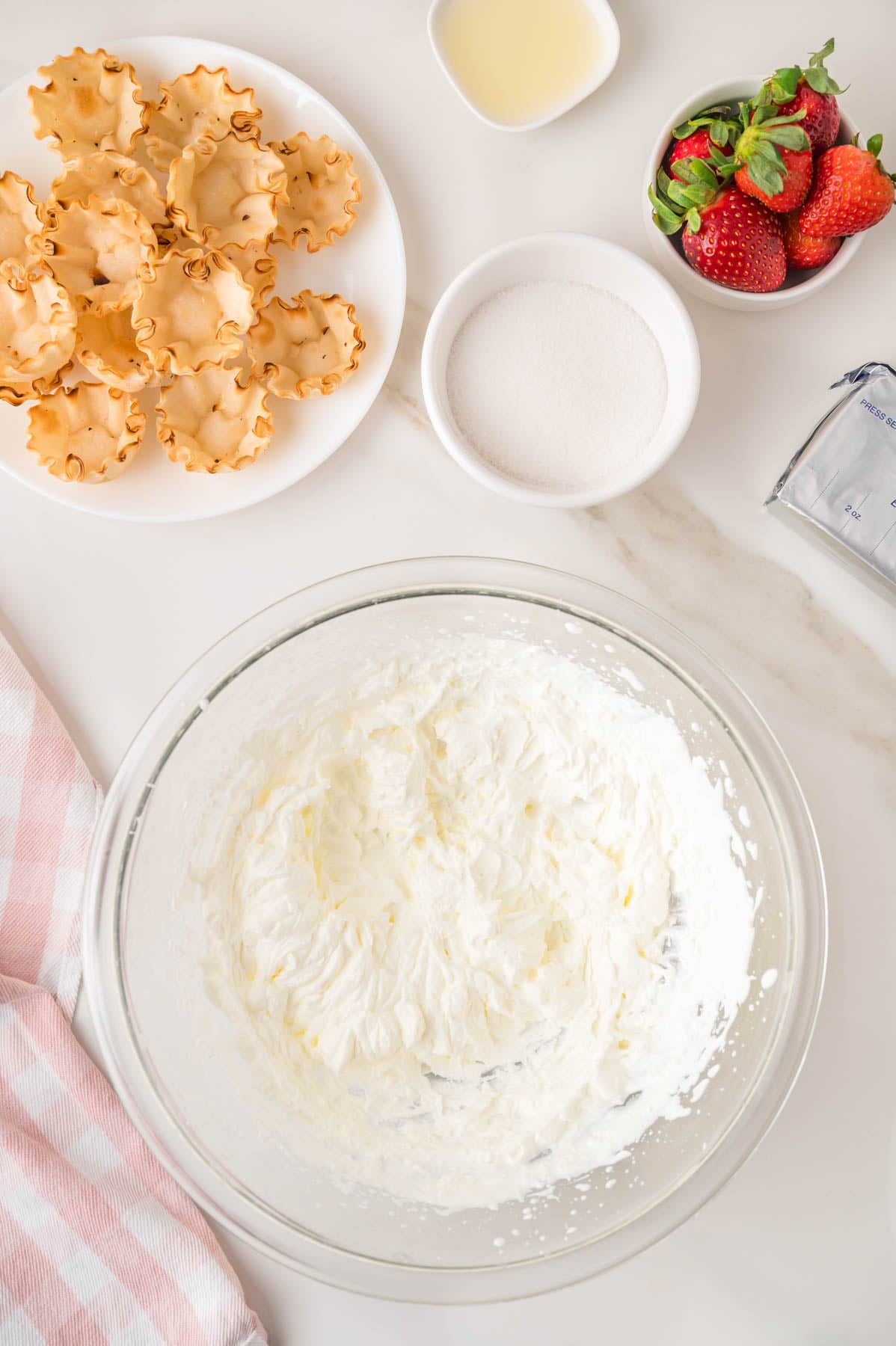 A bowl of whipped cream, sugar, strawberries, pastry cups, cream cheese, and lemon juice sit on a white countertop with a pink-striped towel.