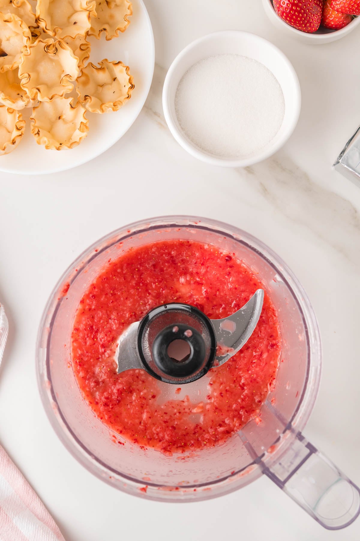 A food processor with blended strawberries sits on a counter near a bowl of sugar, pastry cups, and whole strawberries.