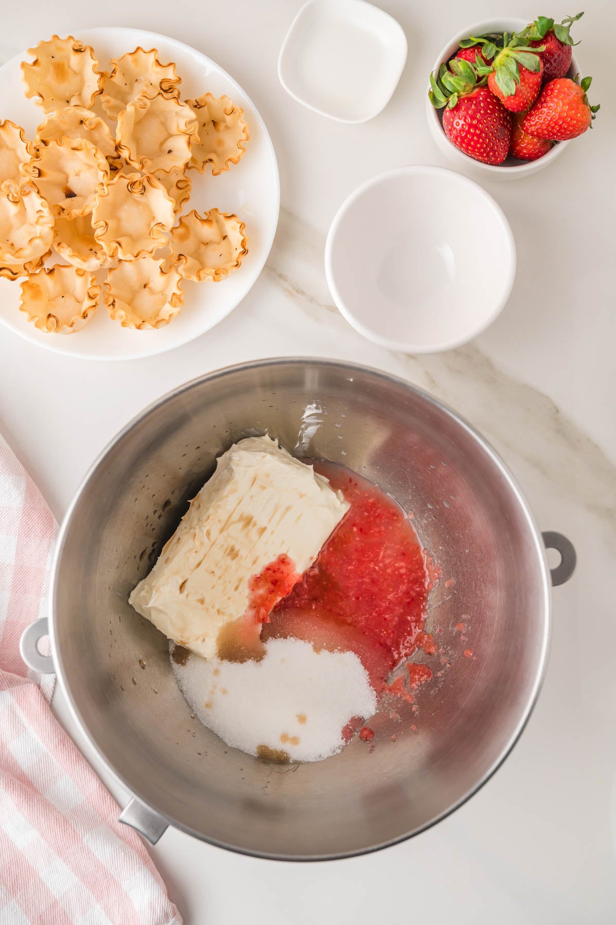 A mixing bowl with cream cheese, sugar, and strawberry puree, next to a plate of pastry shells and a small bowl of strawberries on a marble countertop.