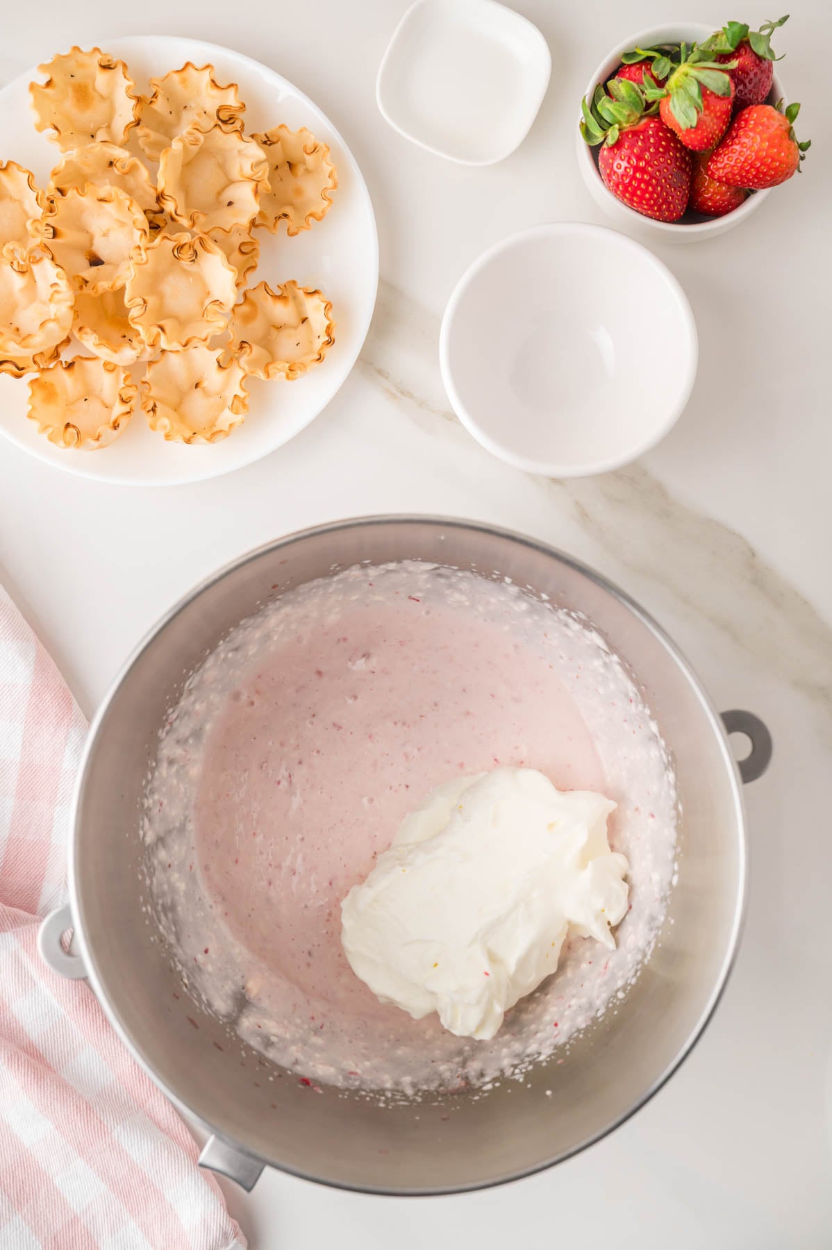 A mixing bowl with pink strawberry cream and a dollop of whipped cream, next to a plate of cookies and a bowl of fresh strawberries on a white counter.