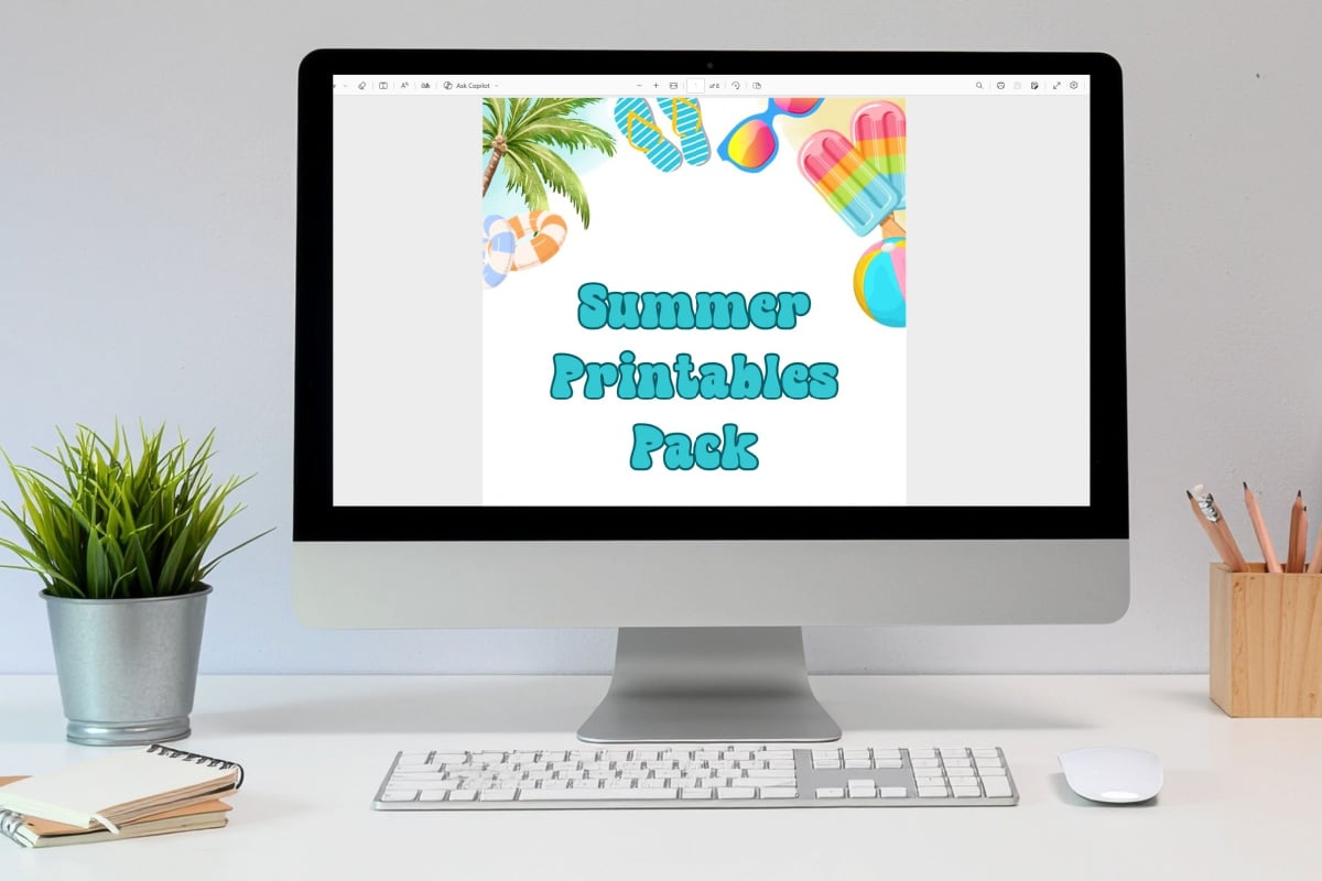 A computer monitor displays a document titled "Summer Printables Pack" with summer-themed graphics. A keyboard, mouse, and potted plant are on the white desk.