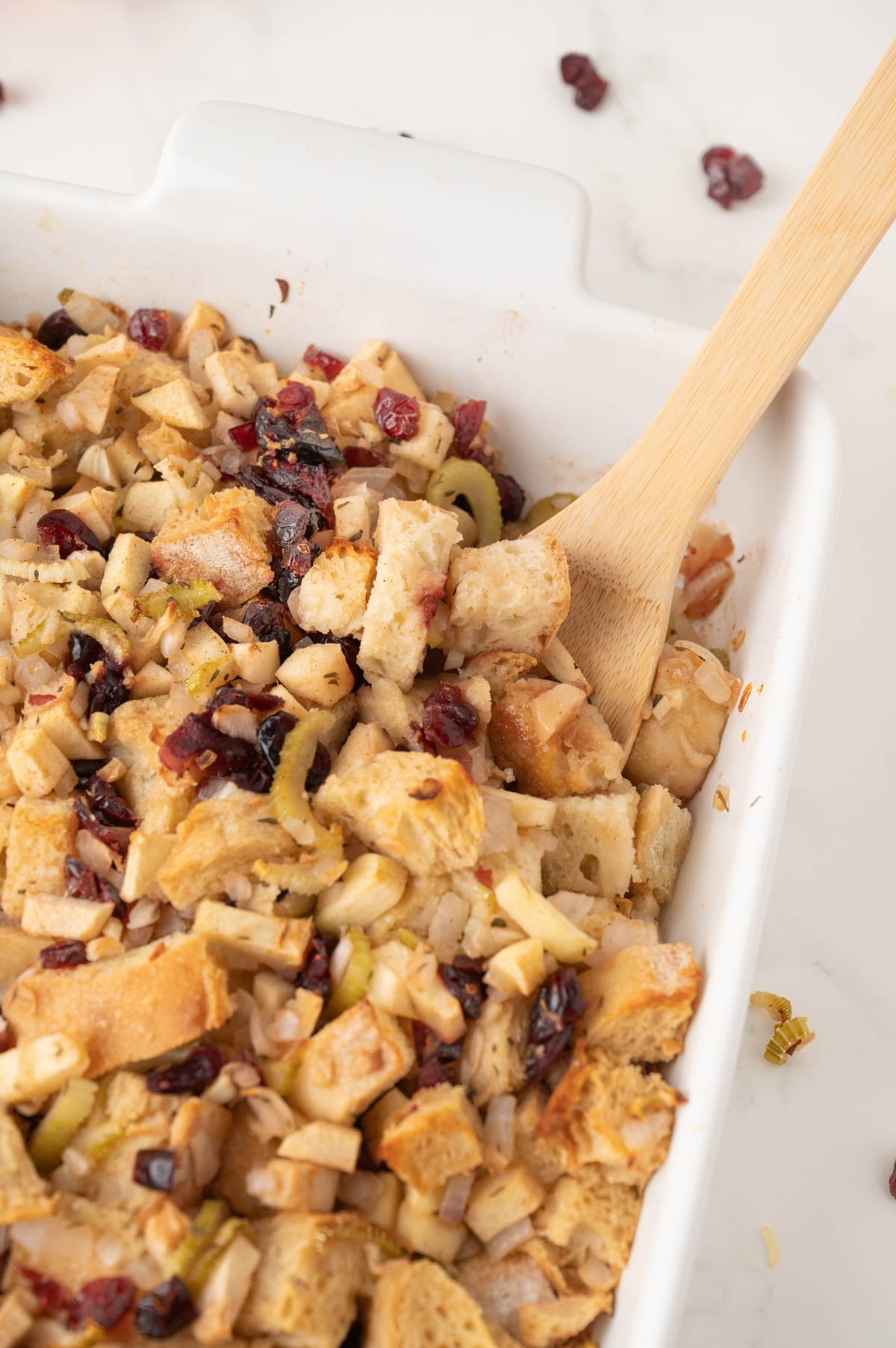 A white baking dish filled with bread stuffing containing chopped apples, celery, and dried cranberries, with a wooden spoon resting inside.