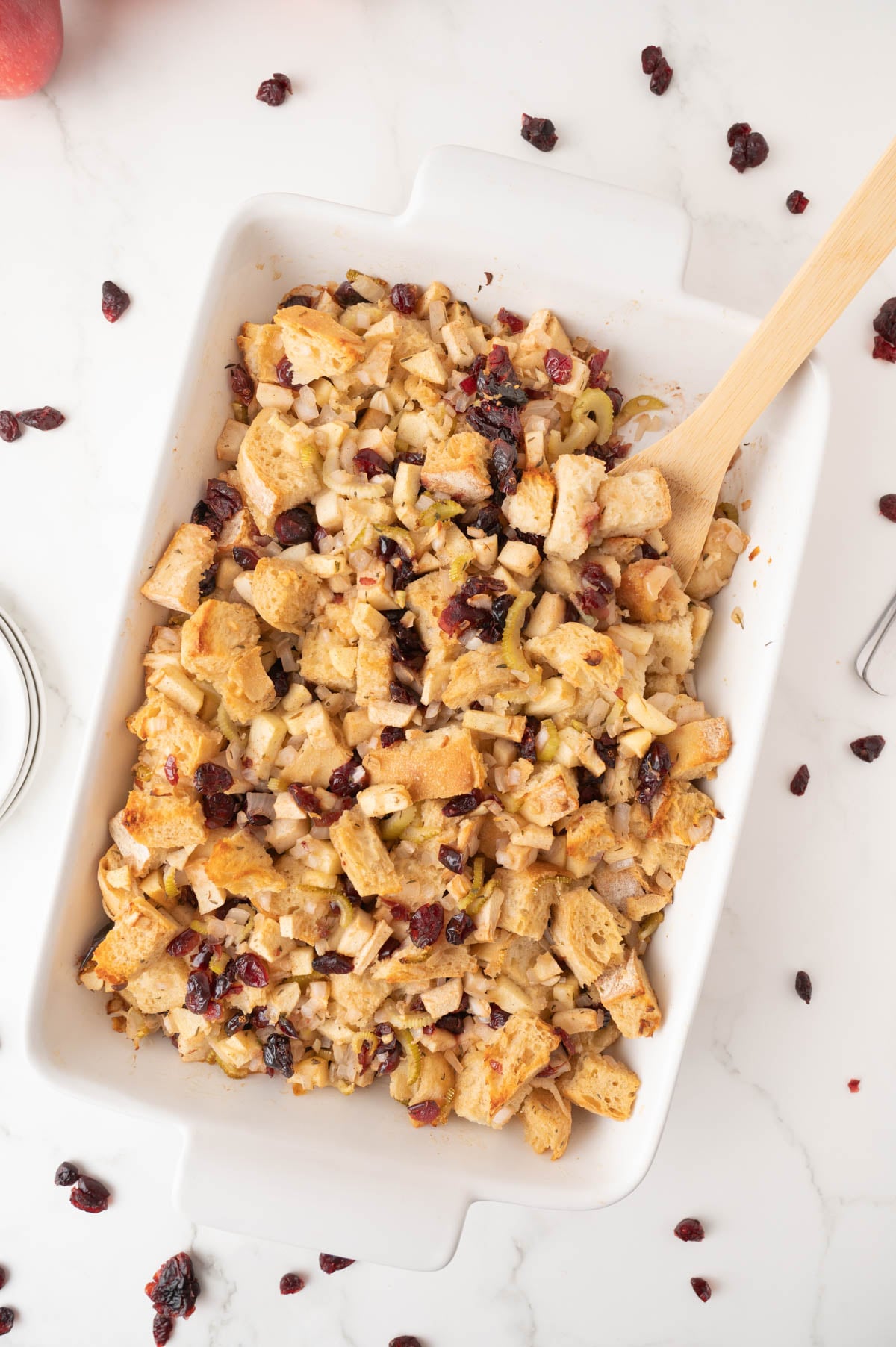A white baking dish filled with bread stuffing mixed with apples and dried cranberries, with a wooden spoon on the side.