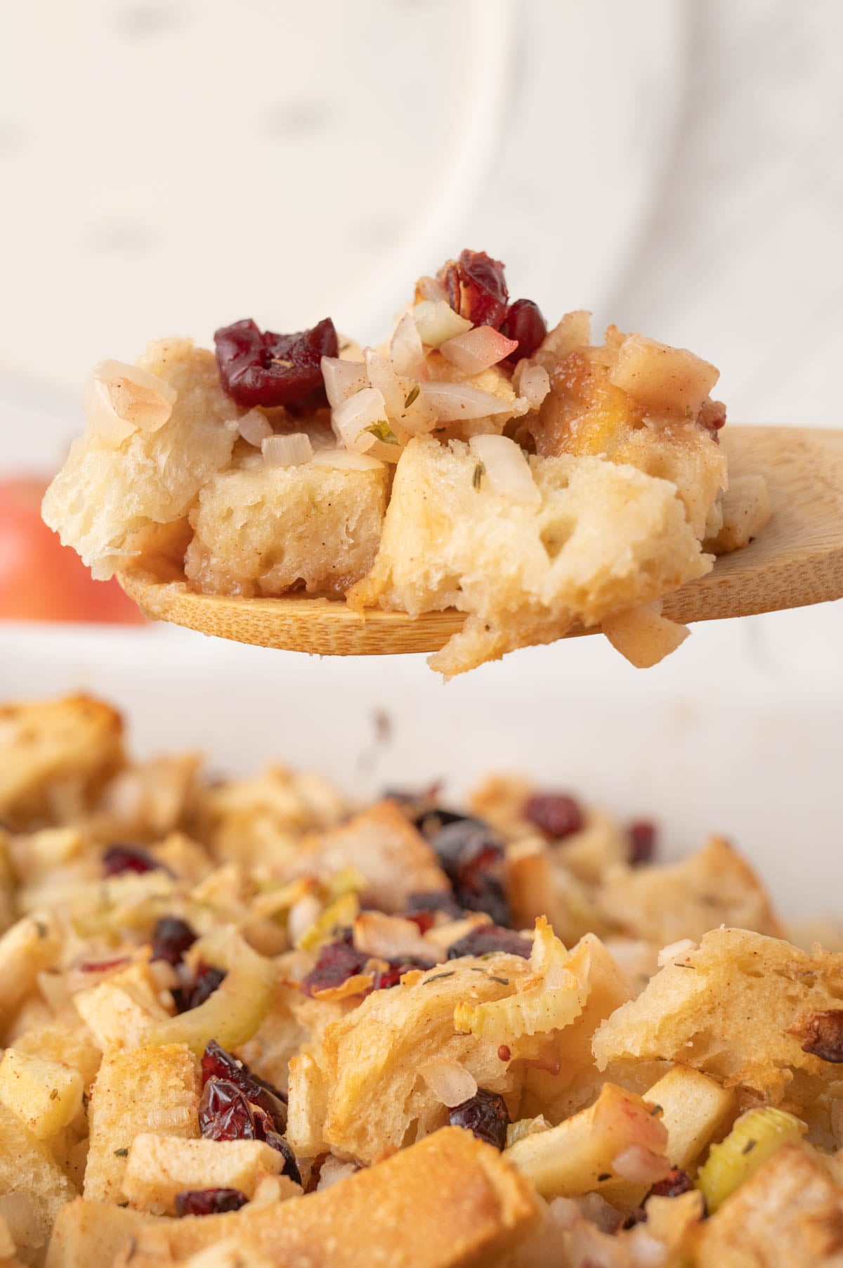 A wooden spoon holds a serving of bread stuffing with chopped onions, celery, and dried cranberries above a dish of the same stuffing.