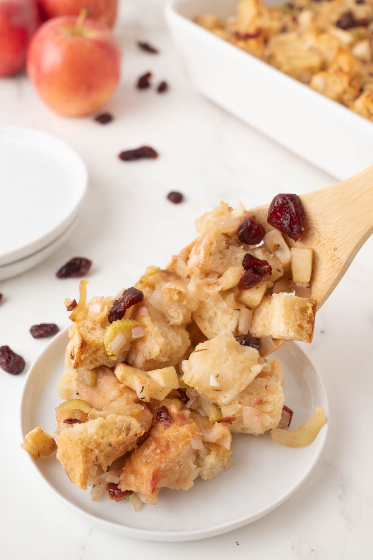 A wooden spoon serves a portion of apple bread pudding with cranberries onto a white plate, with apples and a baking dish in the background.