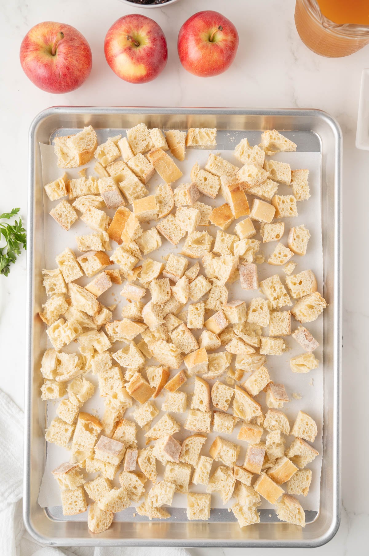 Cubed bread pieces spread on a baking sheet, with apples and a pitcher of cider in the background on a white surface.