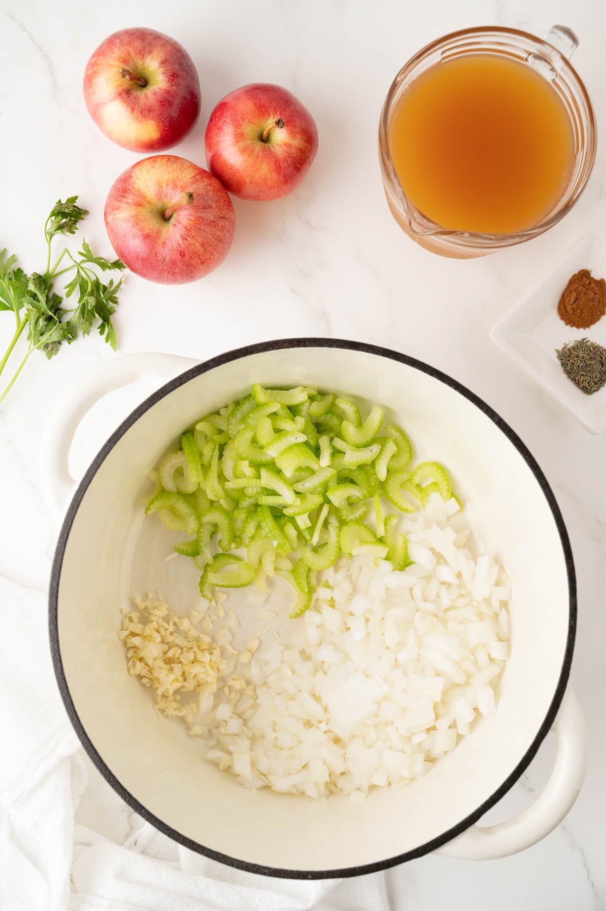 Chopped onions, celery, and garlic in a white pot. Nearby are three apples, parsley, a cup of cider, and a dish with ground spices on a white surface.