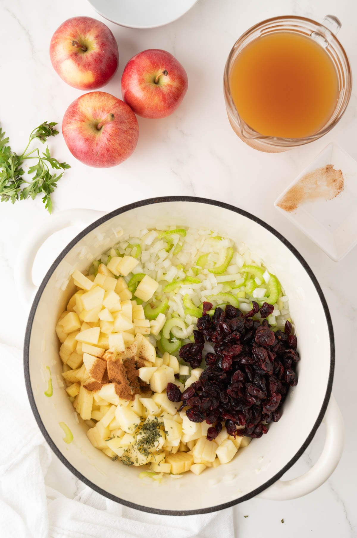 Overhead view of a pot with chopped apples, celery, onions, dried cranberries, and spices. Nearby are three whole apples, a bunch of parsley, a bowl of cider, and a small dish with more spices.