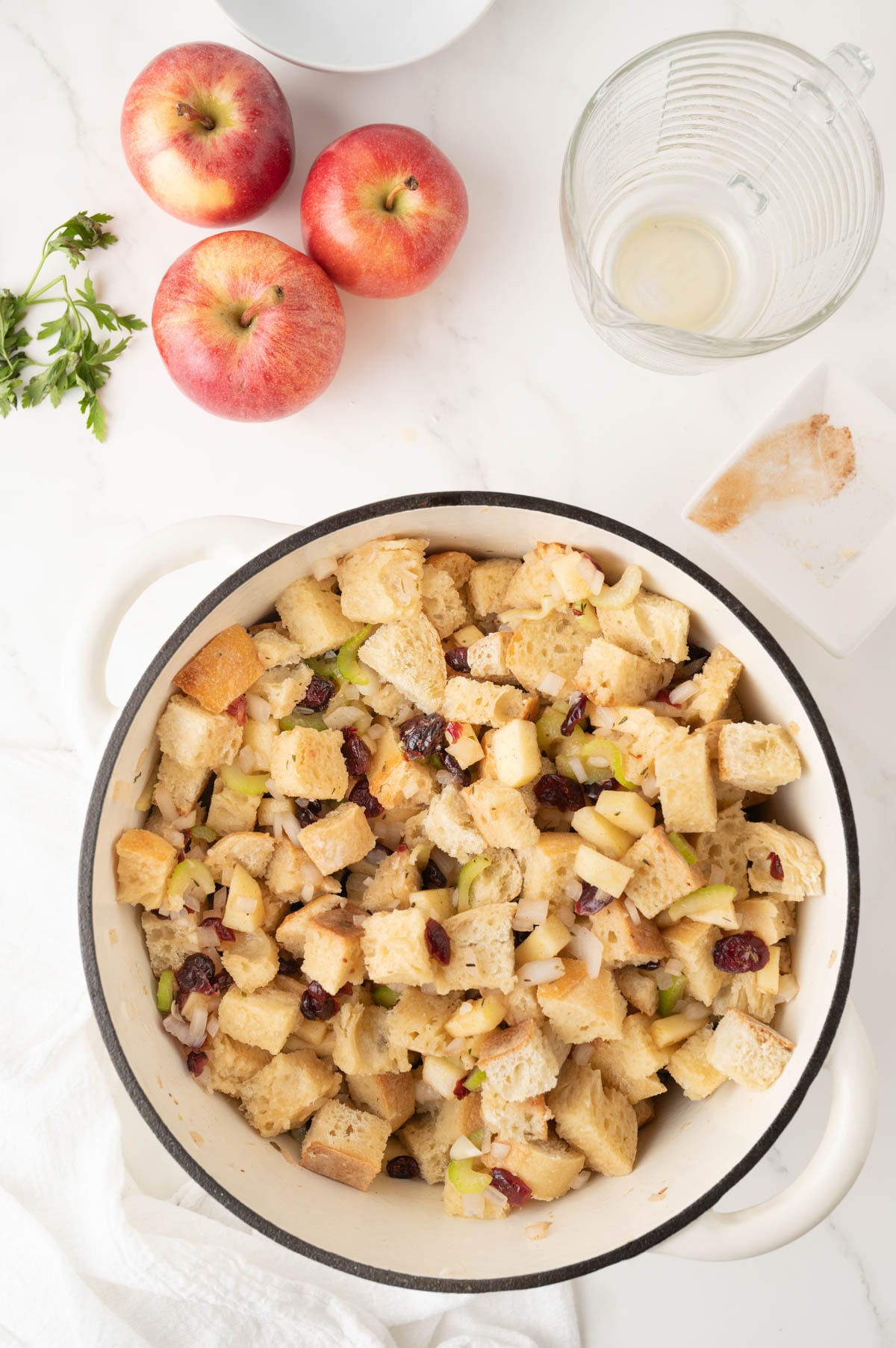 A white pot filled with bread stuffing containing diced apples, celery, onion, and cranberries sits on a marble counter with three apples and an empty glass bowl nearby.