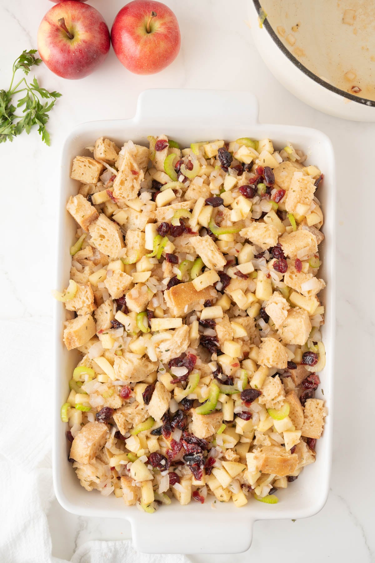 A white baking dish filled with bread stuffing mixed with diced apples, celery, and dried cranberries, on a white countertop with apples and parsley nearby.
