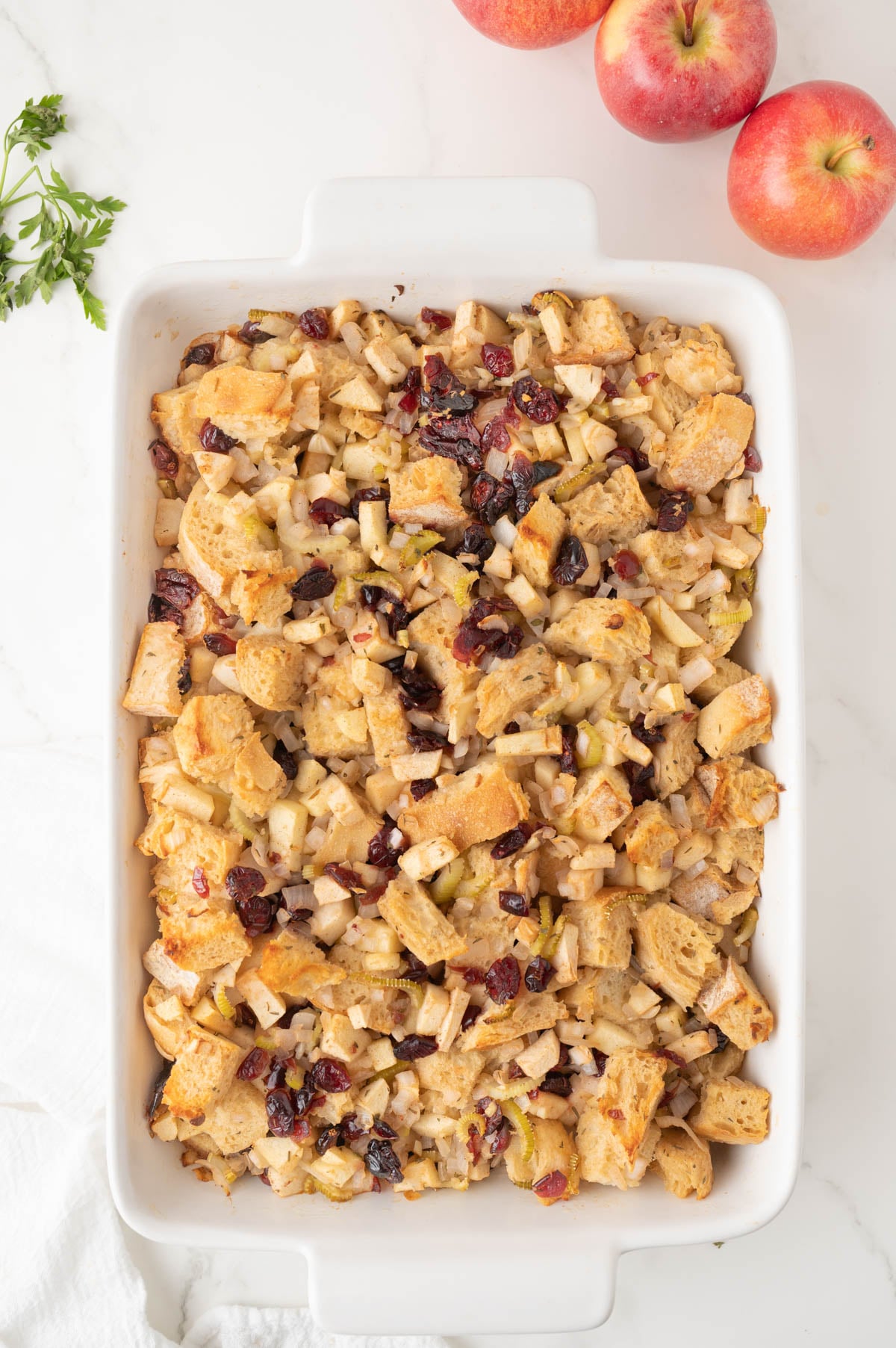 A white baking dish filled with stuffing with apples and cranberries sits on a white surface next to three whole apples and a sprig of parsley.
