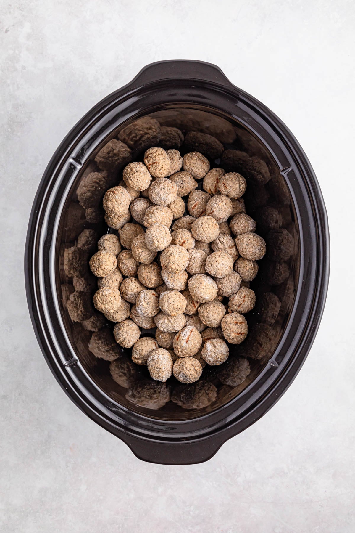 Frozen meatballs arranged in a single layer inside a black slow cooker, viewed from above on a light surface.