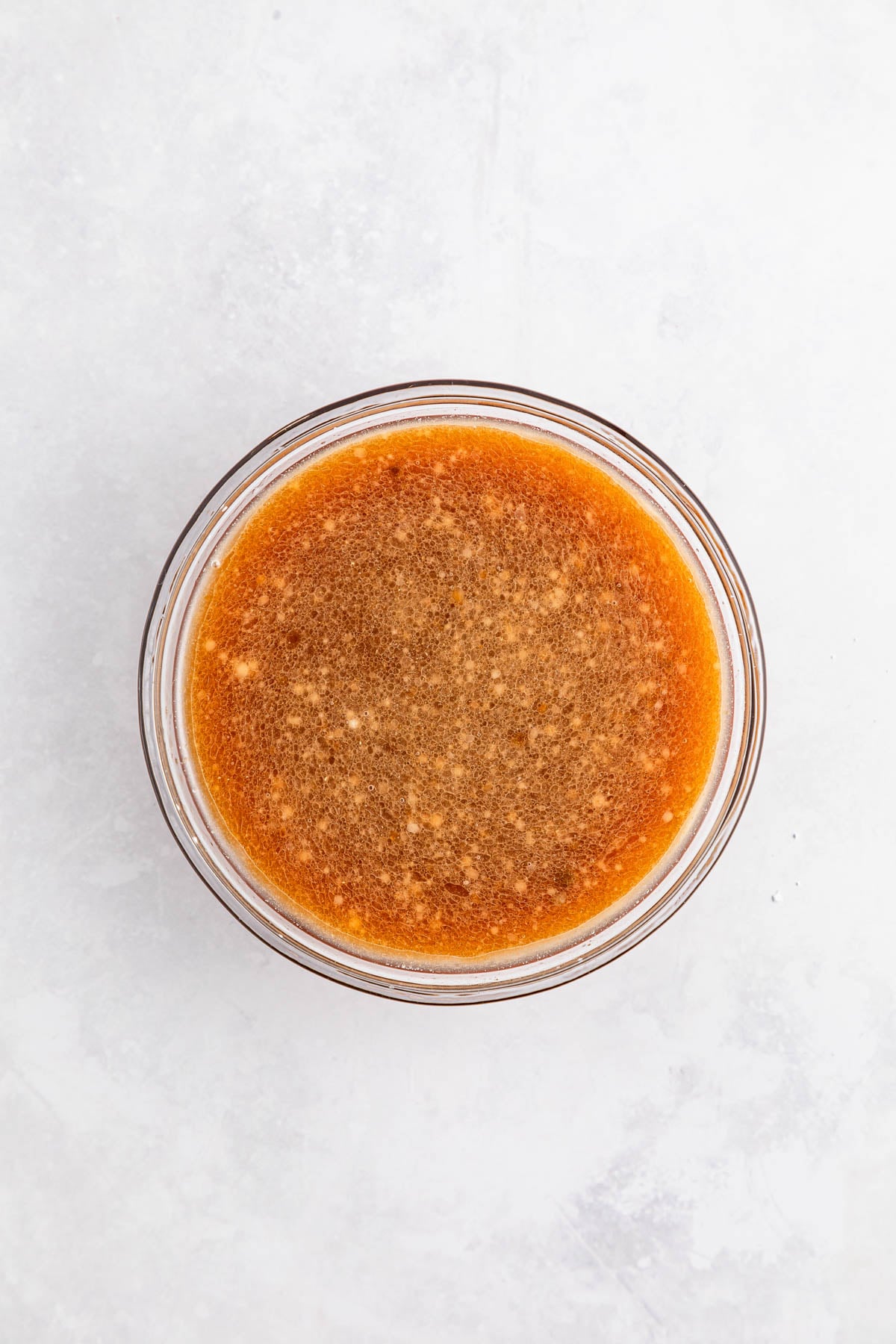 Overhead view of a glass bowl filled with a brown, slightly grainy liquid mixture on a light, textured surface.