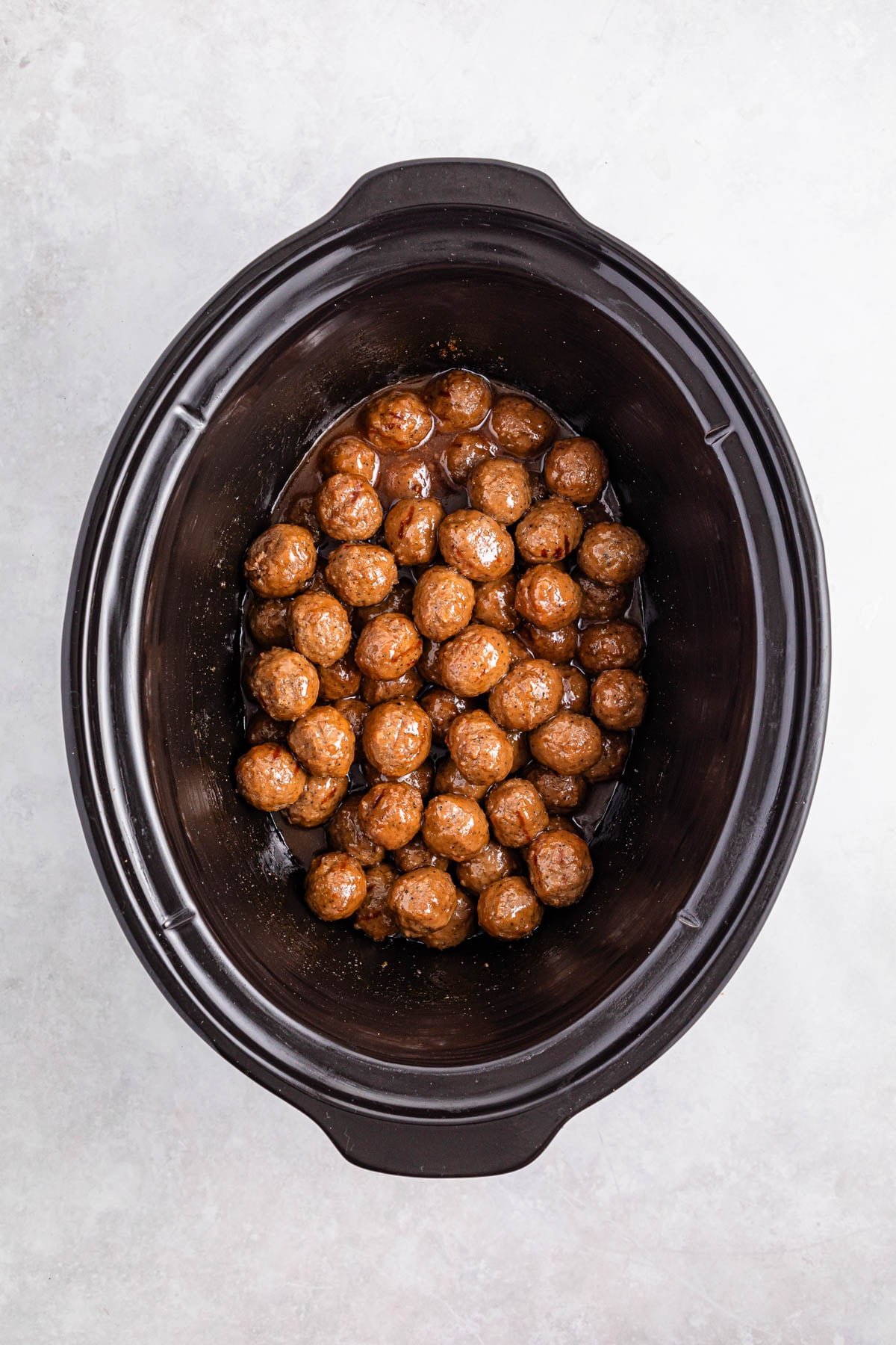 Top view of a black slow cooker filled with cooked meatballs in sauce on a light-colored surface.