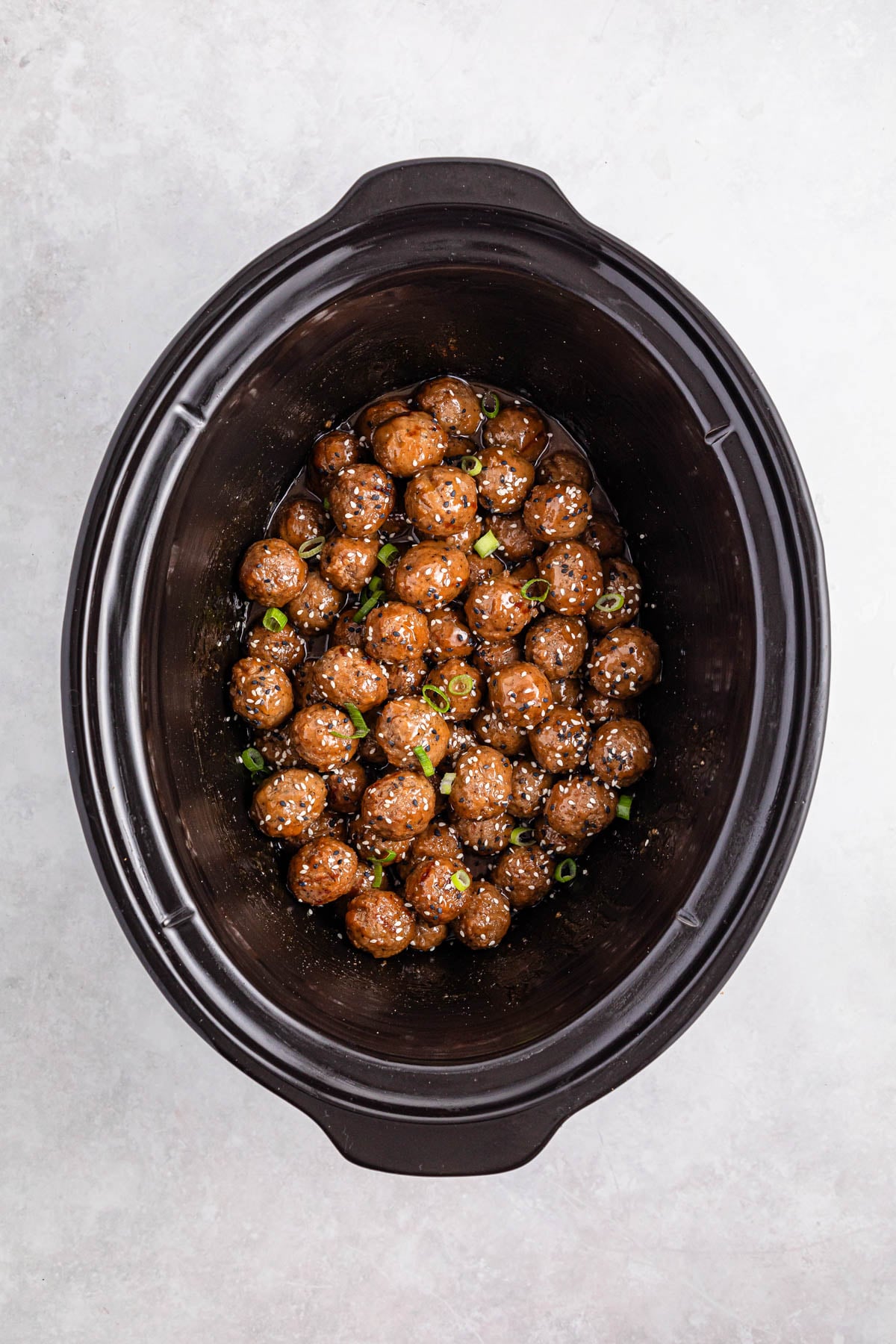 Meatballs coated in sauce are arranged inside a black slow cooker, garnished with sesame seeds and sliced green onions.