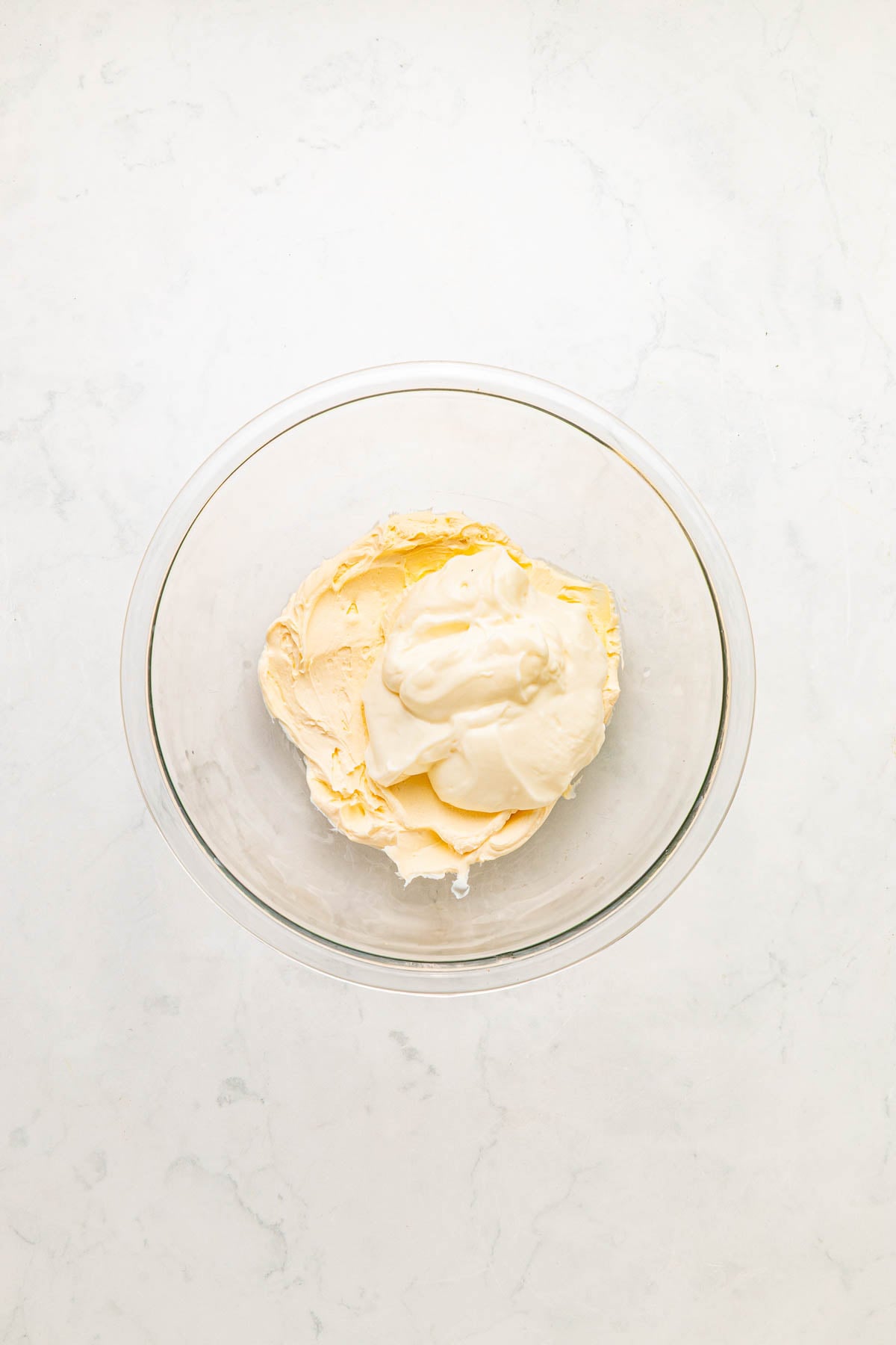 A glass bowl containing a mixture of cream cheese and sour cream on a light marble surface.