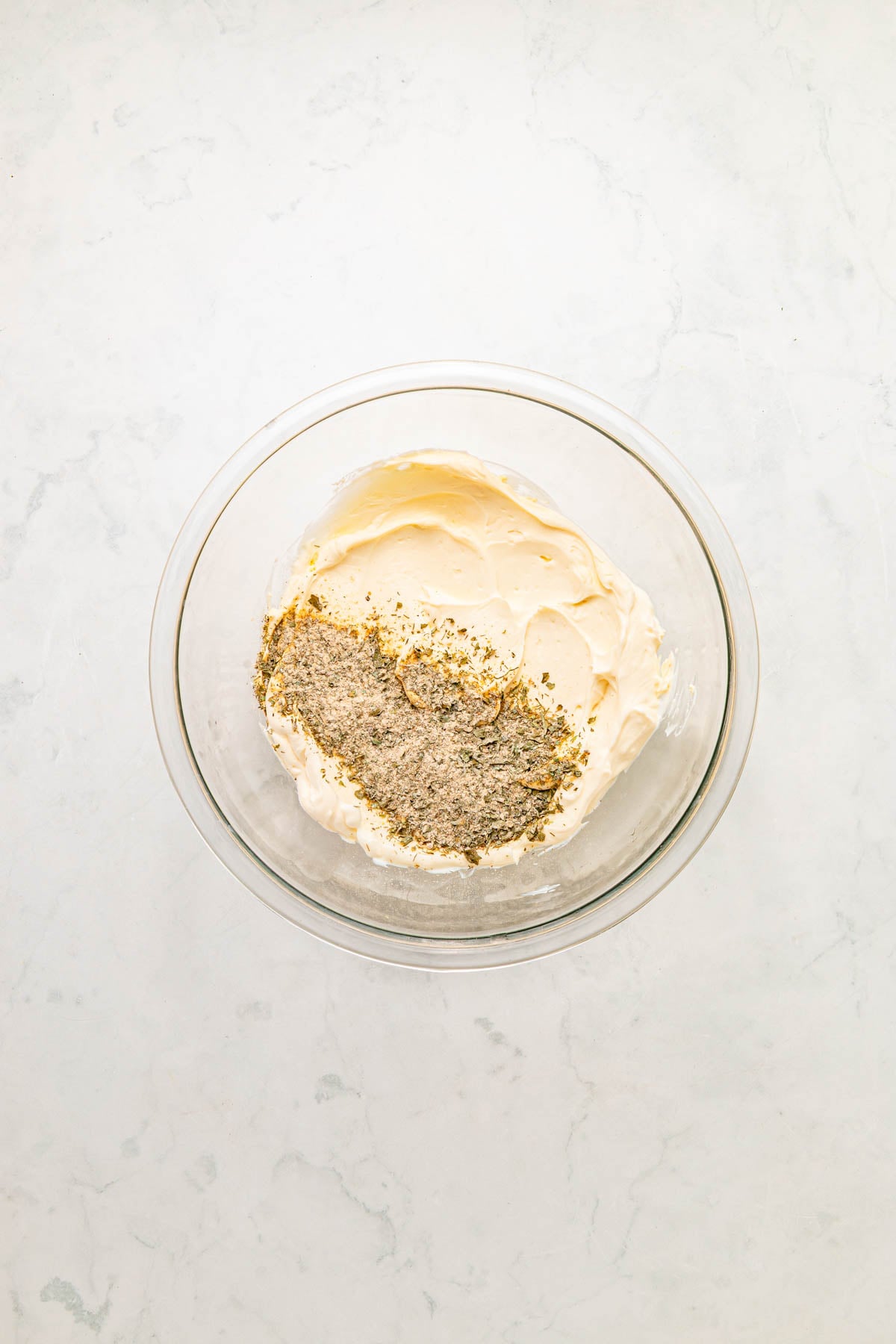 A glass bowl filled with creamy mixture and a layer of herbs and seasoning on top, placed on a light marble surface.