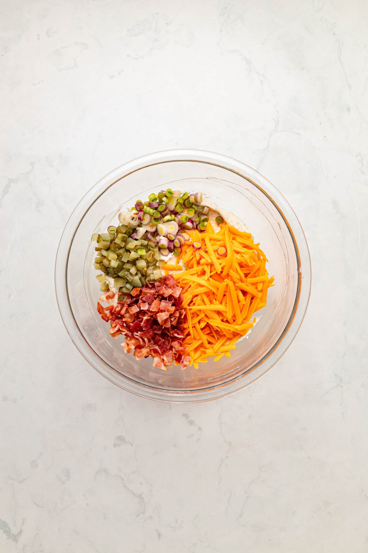 A glass bowl containing chopped bacon, shredded cheddar cheese, diced pickles, chopped red onion, and sliced green onions on a white surface.