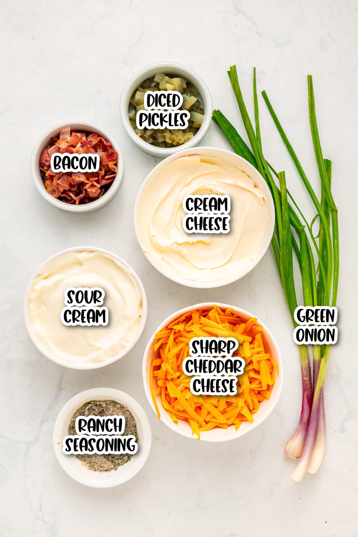 Bowls containing cream cheese, sour cream, ranch seasoning, bacon, diced pickles, and sharp cheddar cheese are arranged on a white surface next to two green onions.