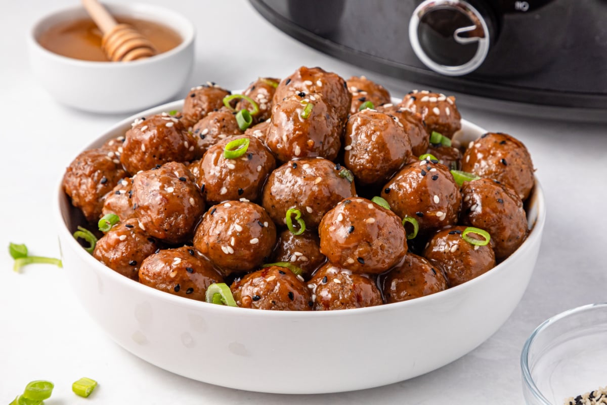 A bowl of glazed meatballs garnished with sesame seeds and chopped green onions sits in front of a slow cooker. Honey and a dipper are visible in the background.